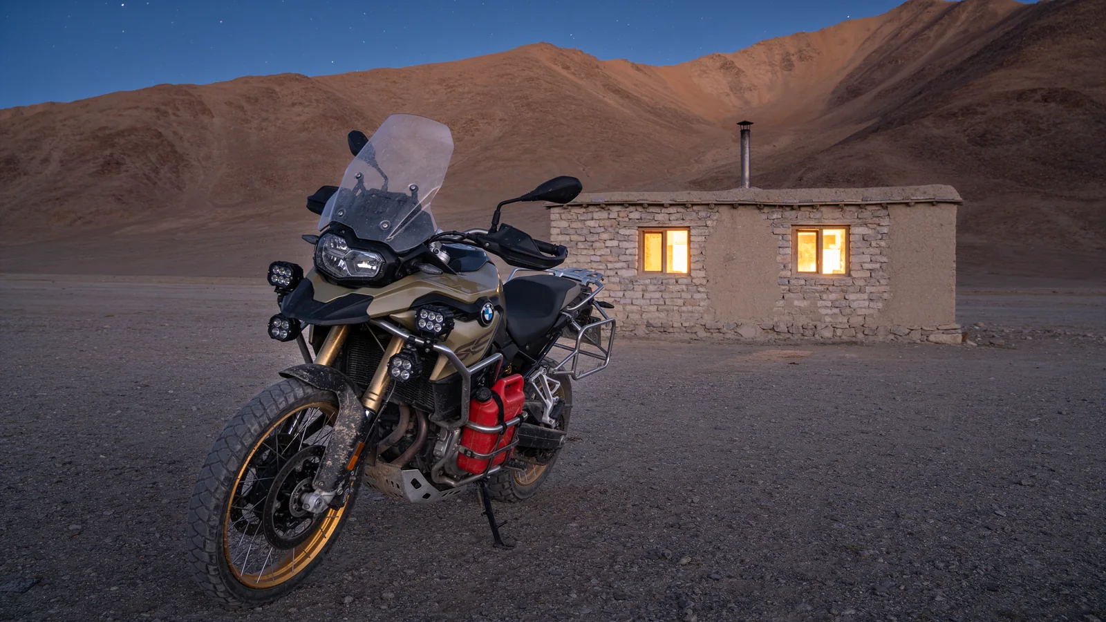 An adventure motorcycle parked for the night outside a small stone guesthouse on a bare high-altitude plateau at dusk, with lit windows and ridgelines behind.